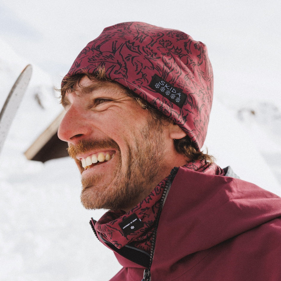 A man smiling outdoors in the snow, wearing a burgundy patterned beanie and a matching burgundy ski jacket with black accents.