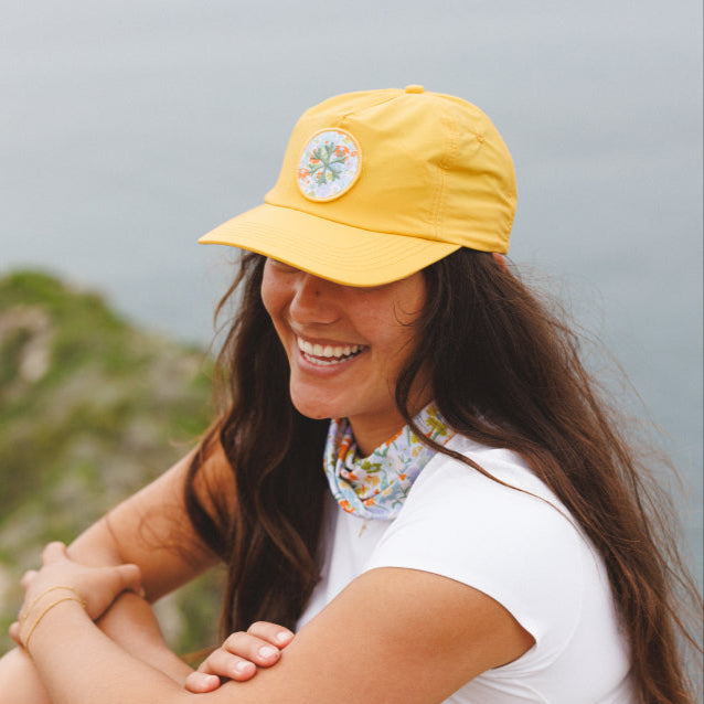 Woman wearing a yellow cap with a logo, sitting outdoors with a scenic background.