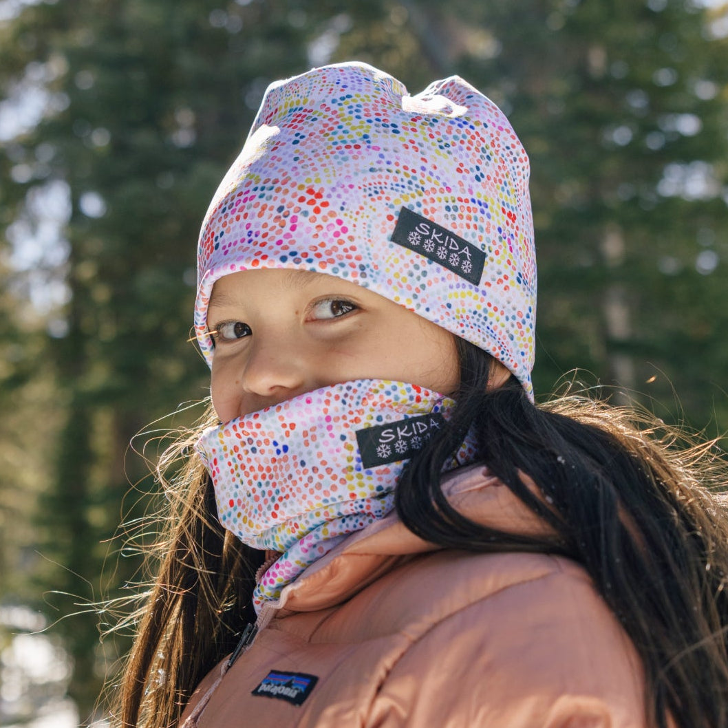 Kid wearing a colorful beanie and face mask with visible branding in a snowy forest setting